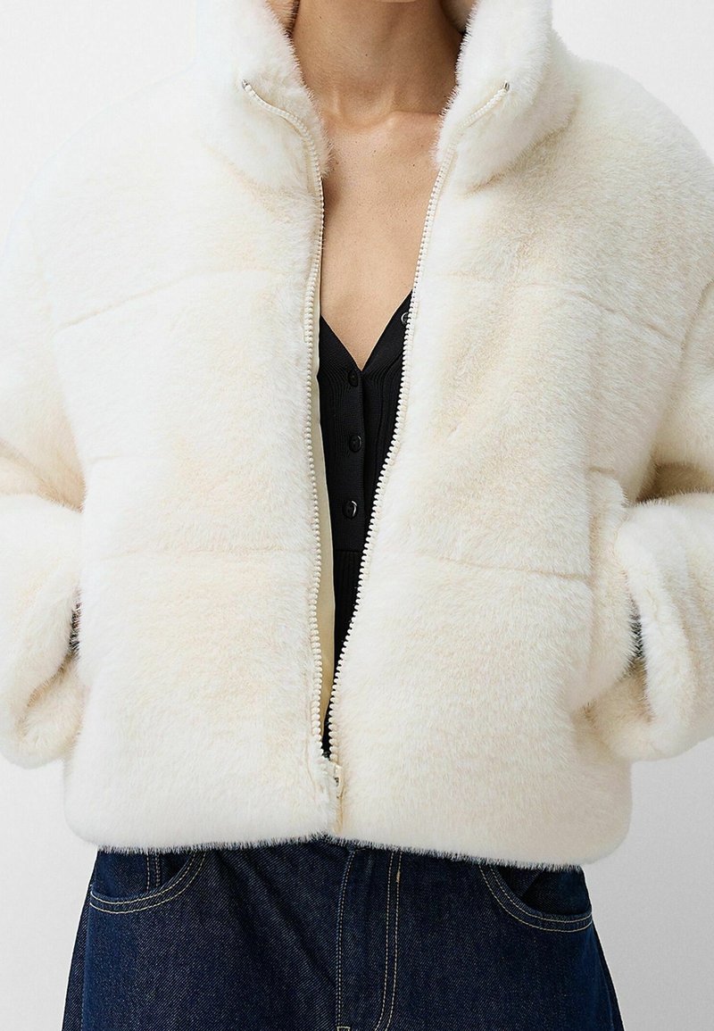 Veste en fausse fourrure couleur crème avec fermeture éclair sur le devant, col montant et poches latérales, présentant une surface douce et texturée et un design court.
