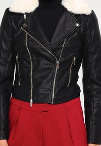 Chaqueta de motocicleta de cuero negro con un collar de piel sintética blanca. Cuenta con cremalleras plateadas y un diseño ajustado, llevada sobre un suéter negro de cuello alto.