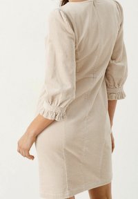 Robe en velours côtelé beige avec des manches ballon trois quarts, des poignets volants et une silhouette ajustée, dotée d'une texture en côtes verticales.