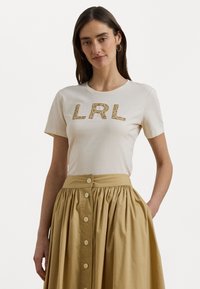 Kreminė trumpomis rankovėmis marškinėliai su tekstūriniu auksiniu "LRL" grafiniu raižiniu. Derinama su aukštuoju juosmeniu, mygtukiniu smėlio spalvos sijonu.
