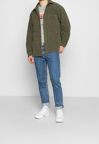 Oliven grøn button-up jakke med en tekstureret overflade, grå t-shirt underneath, blå jeans og hvide sneakers. To frontlommer på jakken.