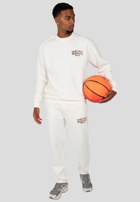 Sudadera blanca y pantalones de chándal con el logotipo "WRONG FRIENDS" en marrón. El modelo sostiene un balón de baloncesto naranja, vistiendo zapatillas gris.