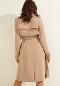 Manteau beige présentant un design croisé, une taille ceinturée et des épaulettes. Texture lisse avec une coupe mi-longue.