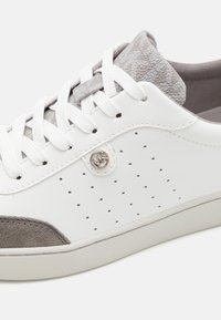 Valkoiset ja harmaat lenkkarit, joissa on reikäkangaspaneli sivussa, valkoiset nauhat, teksturoitu harmaa kieli, mokkanahka kärkipää ja pieni pyöreä metallilogo nauhojen lähellä.