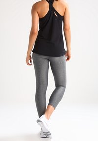 Nainen, joka on pukeutunut mustaan hihattomaan puseroon, harmaisiin leggingseihin ja valkoisiin lenkkareihin, seisoo yksi jalka hieman toisella takana valkoista taustaa vasten.