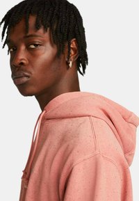 Kurta w kolorze przykurzonego różu z kapturem na sznurki, o teksturowanej tkaninie i cętkowanych detalach. Posiada widoczną przeszywkę oraz boczne szwy.