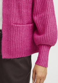 Pink knitted cardigan with ribbed texture, featuring a front pocket and fitted cuffs. Shown worn over a white top and dark pants.