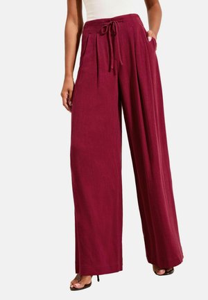 Nő magas derekú, széles szárú bordó nadrágban, elöl pliszézve és derékban megkötve, fehér felsővel és fekete magassarkú szandállal párosítva.