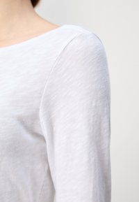 Zbliżenie osoby w białej koszuli z długim rękawem, ukazujące ramię i górną część ramienia na jednolitym tle.