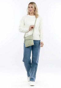 Borsa a tracolla verde, con texture, dotata di due scomparti con cerniera e tracolla regolabile, abbinata a un maglione bianco e jeans blu.