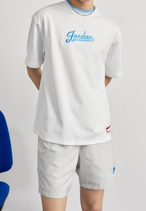 Personne portant un t-shirt Jordan blanc avec un logo bleu et un short gris clair, debout les mains derrière le dos devant un fond uni.