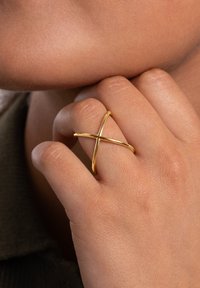 Gouden kruisring met een minimalistisch ontwerp van glad, gepolijst metaal. De band kruist op een centraal punt voor een eigentijdse uitstraling.