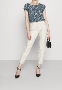 Blouse florale navy avec manches courtes, pantalon crèmes cintrés et escarpins noirs à bout pointu. Petit sac à main noir accentue la tenue.