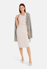 Beige aansluitend jurk gecombineerd met een gestructureerde grijze cardigan. Glanzende zilveren pumps met spitze neus maken de look compleet.