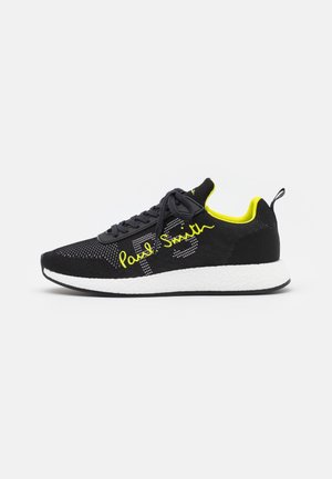 Schwarz-gelber Sportschuh mit weißer Sohle, gestricktem Obermaterial, Schnürung vorne und "Paul Smith" Text an der Seite.