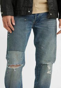 Jeans de mezclilla azul con detalles desgastados, que presentan adornos plateados y un desgarro en la rodilla. Diseñados con un ajuste holgado.