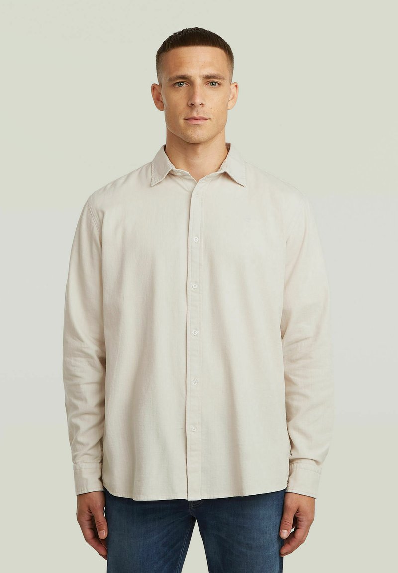 Mann steht mit dem Gesicht nach vorne, trägt ein beigefarbenes langärmliges Hemd mit Knöpfen und blaue Jeans vor einem einfachen hellen Hintergrund.