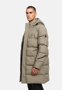 Pūkuotas paltas šviesiai beige spalvos, su gaubtu, užtrauktukais kišenėse ir logotipo detalėmis. Dizainas apima siuvinėtą tekstūrą ir ilgas kelnes.