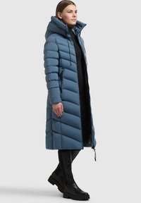 Ilgs zils pūkainais mētelis ar kapuci, ar ševrona rakstu, ar rāvējslēdzēju priekšpusē un sānu kabatām. Valkāts ar augstām melnām zābakiem līdz ceļgaliem.