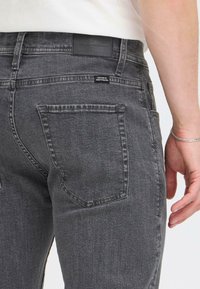 Grijze denim jeans met een slanke pasvorm, voorzien van een zichtbaar zakje en een zwarte leren taillebandlabel. Subtiele textuur en klassieke stiksels.