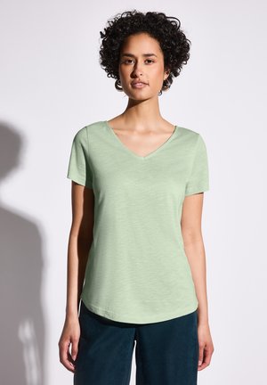 T-shirt vert clair en v, fabriqué en tissu doux et texturé. Manches courtes et ourlet arrondi. Porté avec un pantalon bleu foncé.