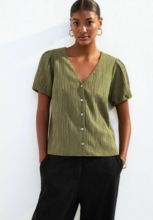 Olijfgroen, textuur blouse met een V-hals, korte puffmouwen en sluiting met knopen aan de voorkant. Gecombineerd met zwarte high-waisted broek.
