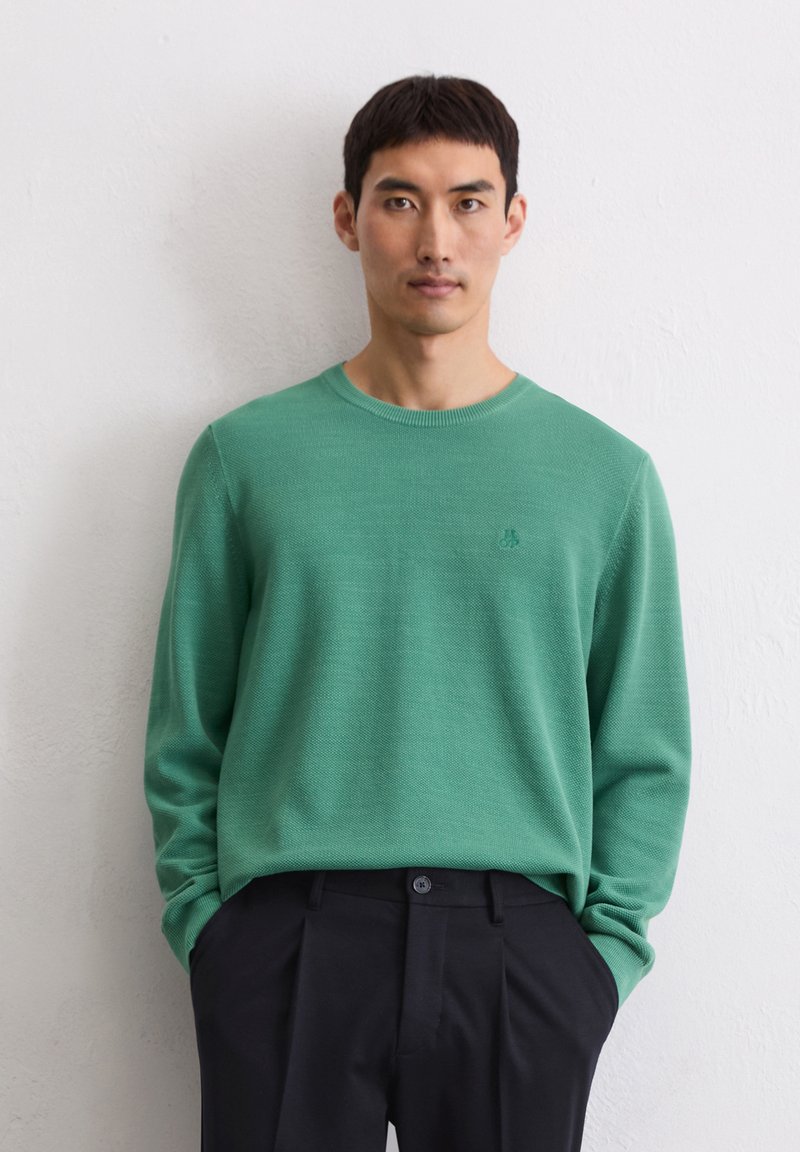 Maglione verde a maglia testurizzata con scollo rotondo e maniche lunghe, abbinato a pantaloni plissettati neri. Dettaglio del logo discreto sul petto.