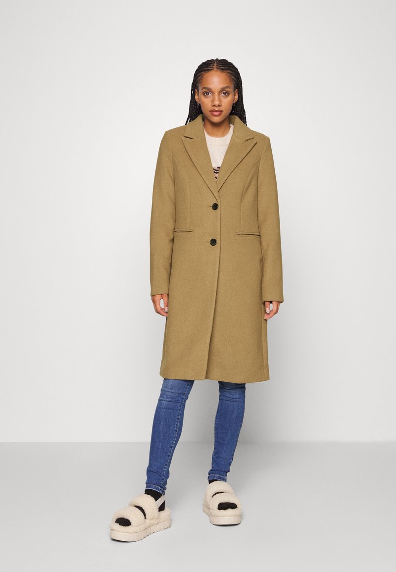 Beige ullblandet frakk med klippte revers, frontlukk og sidelommer, kombinert med blå jeans og fluffy beige platåsko.