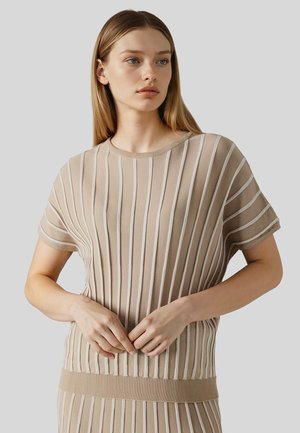 Młoda kobieta z prostymi blond włosami, ubrana w beżową bluzkę z krótkim rękawem i pionowymi białymi paskami, stojąca na szarym tle.