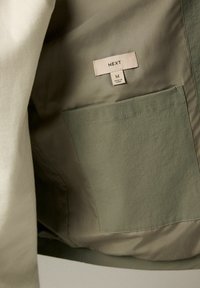 Lyse beige beklædning indeni med en grøn lomme, stofetiket mærket "NEXT," størrelse "M," og "MADE IN CHINA." Glat tekstur, enkelt design.