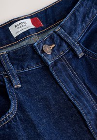 Jean en denim bleu foncé à taille mi-haute, étiquette, bouton en cuivre et surpiqûres orange visibles sur la ceinture et les passants de ceinture.