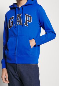 Sweat à capuche zippé bleu en coton. Comprend une poche kangourou et une capuche avec cordon de serrage. Logo "GAP" en blanc avec contour bleu marine sur le devant.