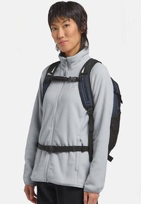 Giacca in pile grigio chiaro con collo alto, cerniera frontale e cinghie regolabili. Zaino nero e blu navy con dettagli in rete.