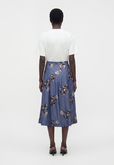 Femme debout, de dos, portant un haut blanc à manches courtes, une jupe midi plissée bleue à motif floral et des sandales noires à talons.