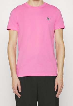 Homme portant un t-shirt rose vif à manches courtes avec un petit logo zèbre sur la poitrine gauche et un pantalon noir.