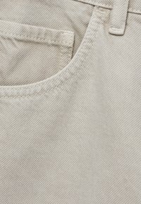 Detalle en primer plano de una tela de mezclilla beige que muestra el bolsillo frontal, el lazo del cinturón y las costuras detalladas en los pantalones.