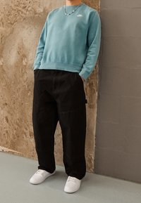 Světlě modrá mikina s kulatým výstřihem a malým logem, v kombinaci s černými volnými kalhotami a bílými teniskami, stojící před strukturovanou zdí.