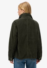 Giacca di velluto a coste verde scuro con collo alto, caratterizzata da un taglio dritto e polsini con bottoni, indossata sopra jeans blu.