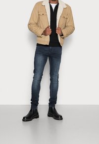 Giacca color marrone chiaro con colletto in shearling bianco, indossata sopra un maglione nero, abbinata a jeans blu scuro e stivaletti Chelsea neri. Sfondo uniforme.