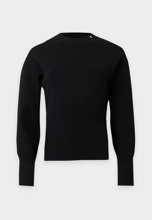 Jeune personne portant un haut noir à manches longues, un jean large bleu et des chaussures pointues marron, debout devant un fond uni.