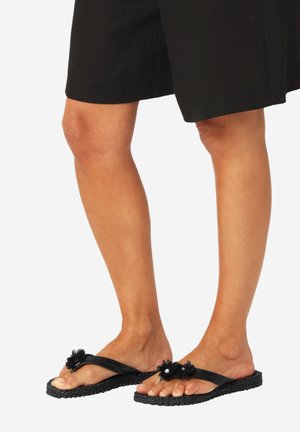 Benen met een zwarte rok tot op de knie en zwarte teenslippers met decoratieve zwarte bloemen, staand op een witte achtergrond.