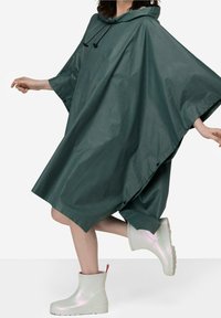 Grøn vandtæt poncho med hætte, der har en løs pasform og sidesnapper, parret med hvide iriserende ankelsko med en rød strop.