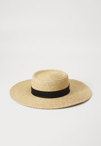 Bred dug straw-hatt med bred brem, flat krone, naturlig farge og et kontrasterende svart stoffband rundt basen. Teksturert vevd design.