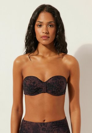 Frau mit mittlerem Hautton und langen, welligen, dunklen Haaren trägt ein trägerloses Bikinioberteil mit dunklem Leopardenmuster vor einem schlichten hellen Hintergrund.