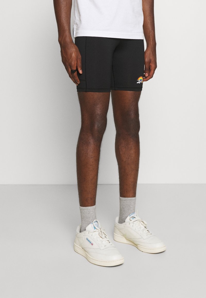 Sorte atletiske shorts med en tætsiddende pasform, prydet med et farverigt regnbue-logo på venstre ben og sammen med hvide sneakers og grå sokker.