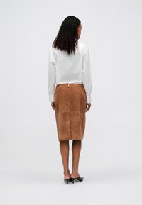 Vrouw met krullend haar, gekleed in een witte blouse met lange mouwen, een bruine suède rok tot op de knie met een rits aan de achterkant en zwarte gespte muilen, rechtopstaand met de rug naar de camera gekeerd.
