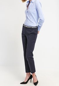 Camicia azzurro chiaro con bottoni, pantaloni tapered blu navy, cintura argentata e tacchi alti neri. Tessuto liscio con vestibilità sartoriale e design minimalista.
