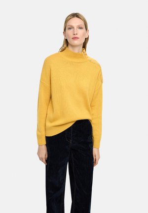 Kobieta stojąca, ubrana w musztardowy golf z bocznym zamkiem błyskawicznym, w połączeniu z luźnymi czarnymi spodniami, na jasnym, jednolitym tle.