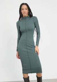 Ribbed, dark green midi dress with long sleeves, high neck, and scalloped hem. Features a central seam down the front and textured fabric.