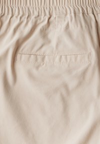Tissu beige clair avec une texture douce. Présente une poche arrière soigneusement cousue avec une ouverture rectangulaire. Ceinture élastique en haut.